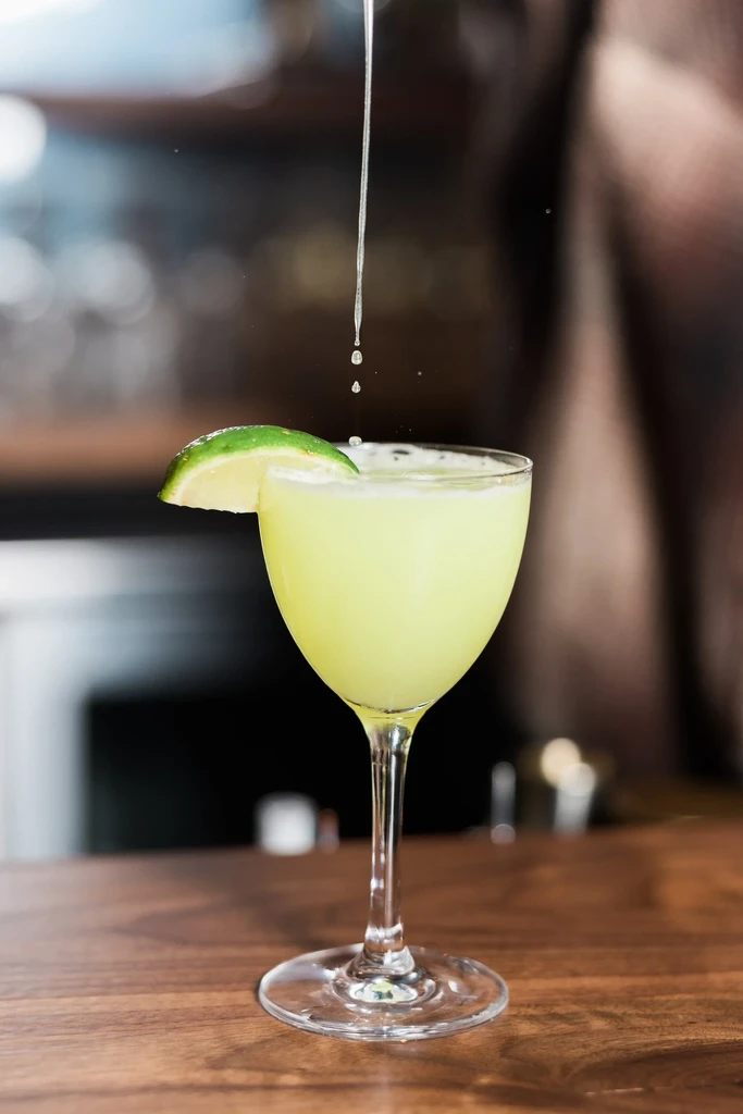 Pale green cocktail in stemmed glass with lime, liquid pouring.