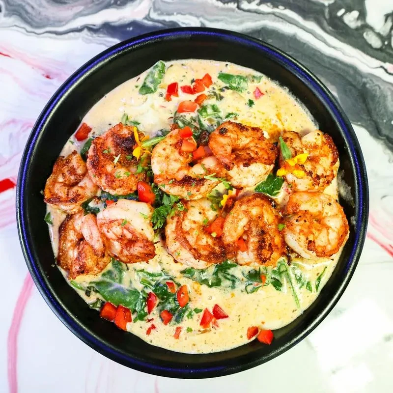 Charred shrimp, creamy sauce, vegetables, and flowers in a dark bowl.