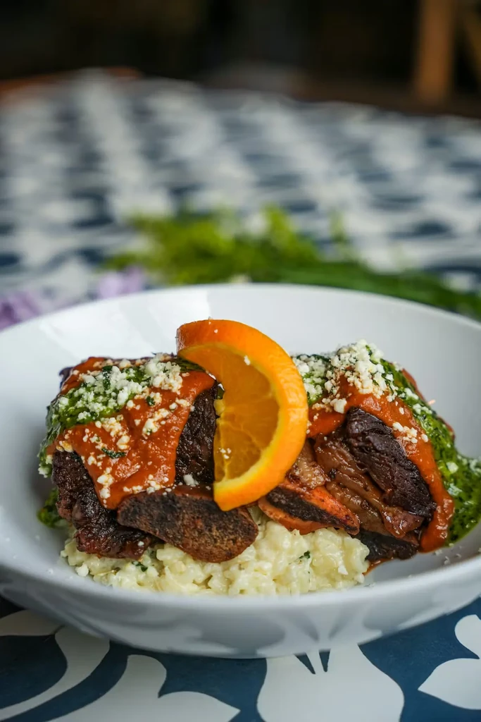 Dark meat dish with red/green sauces, cheese, orange slice, and rice.