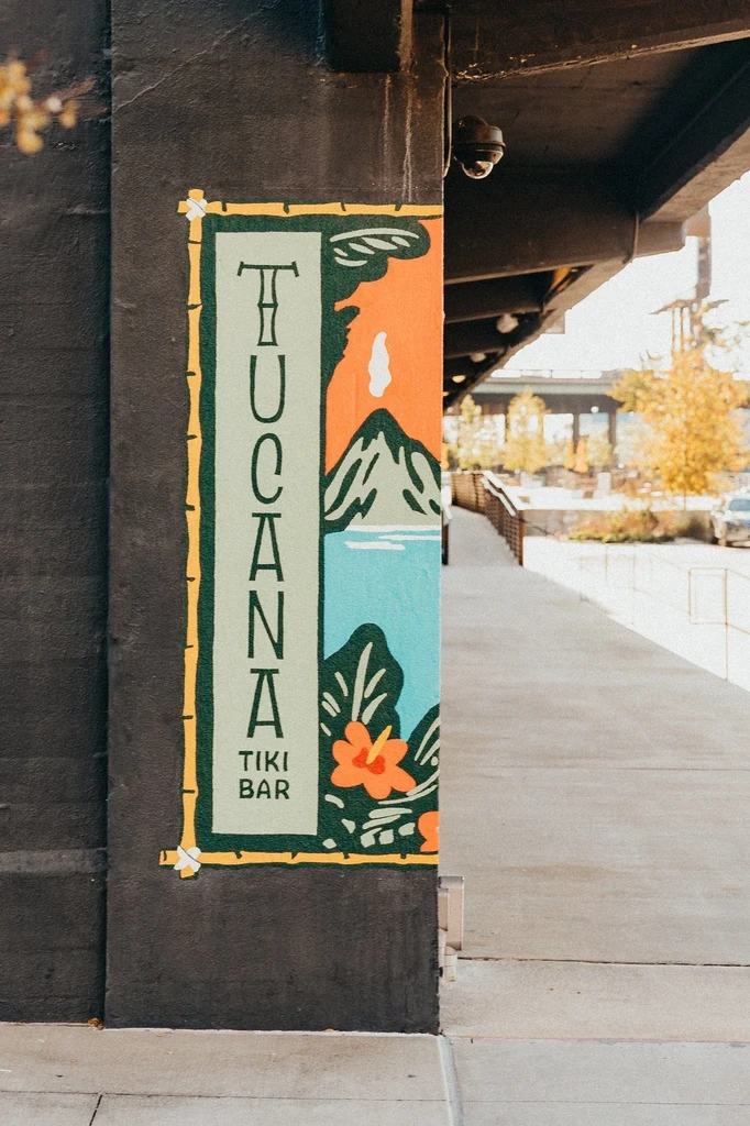 Colorful mural: 'TUCANA TIKI BAR' tropical volcano scene with bamboo frame.