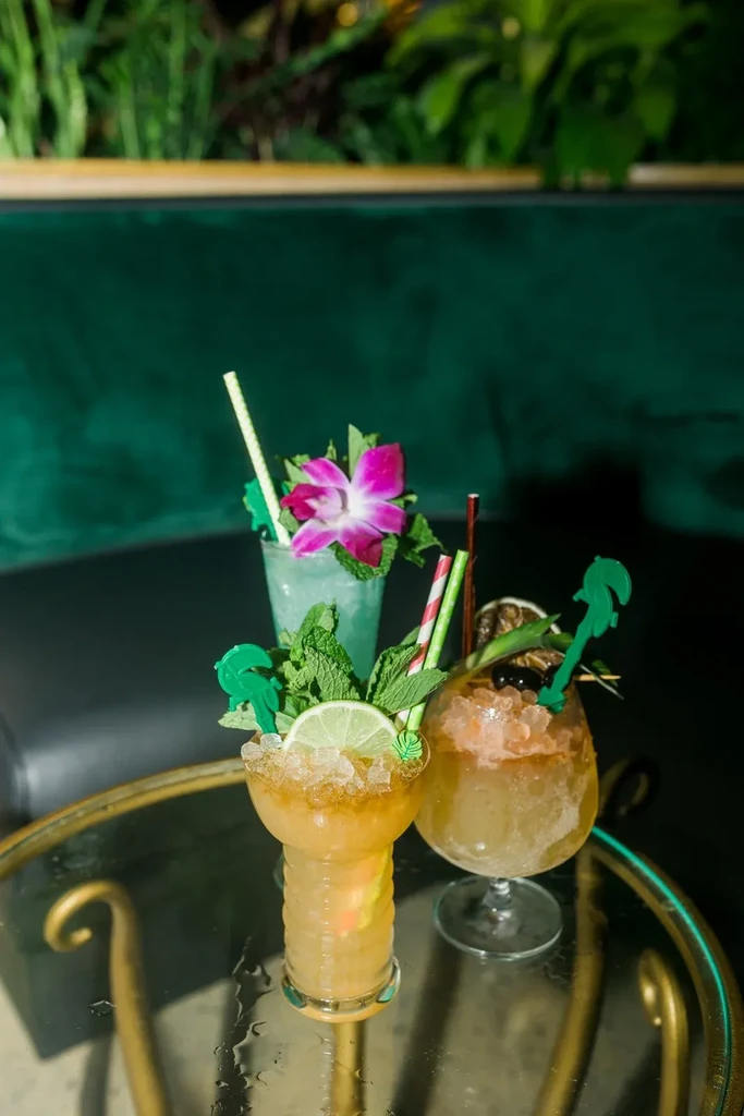 Three elaborately garnished cocktails on a glass table with tropical decor.