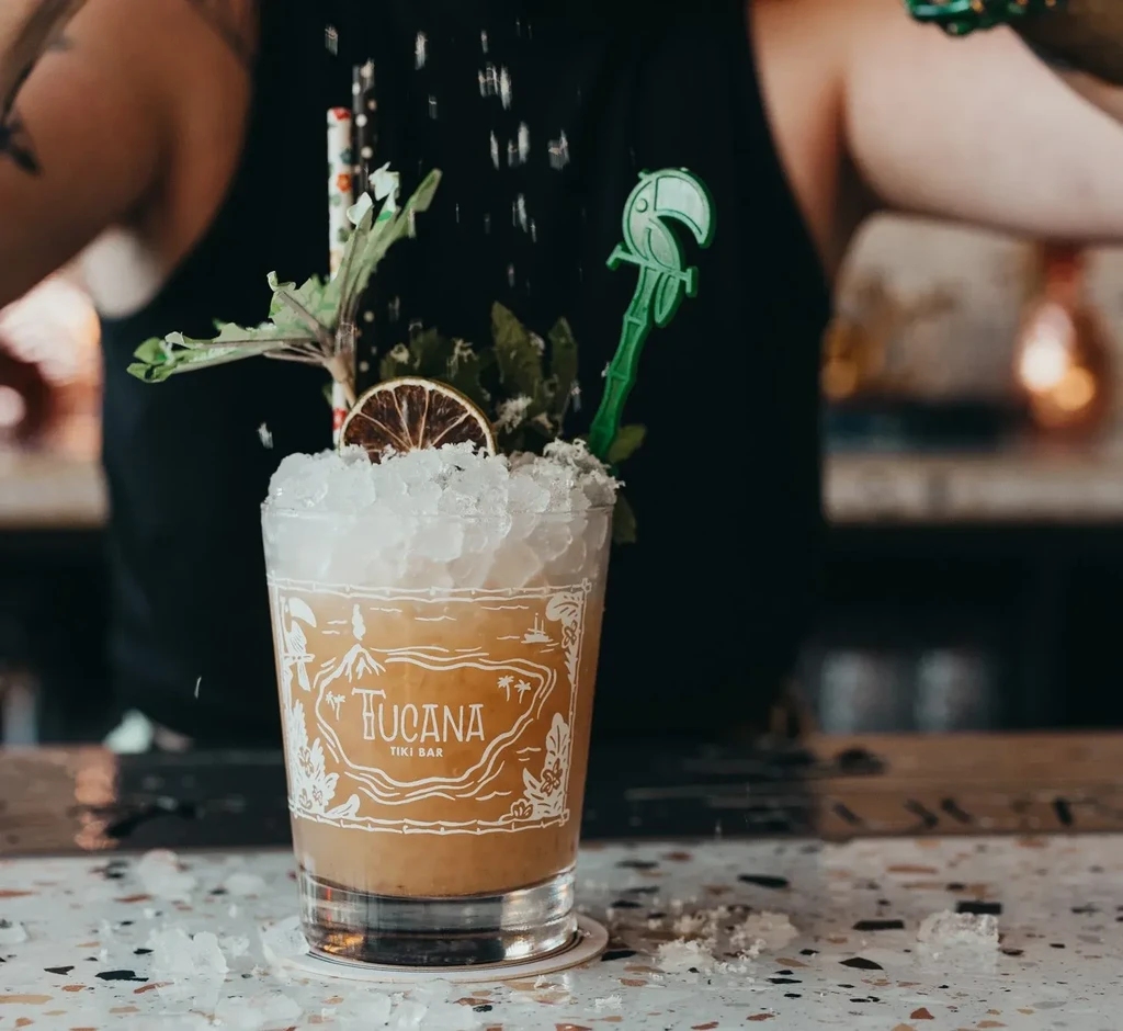Bartender preparing a garnished tiki cocktail with crushed ice, toucan stir stick.