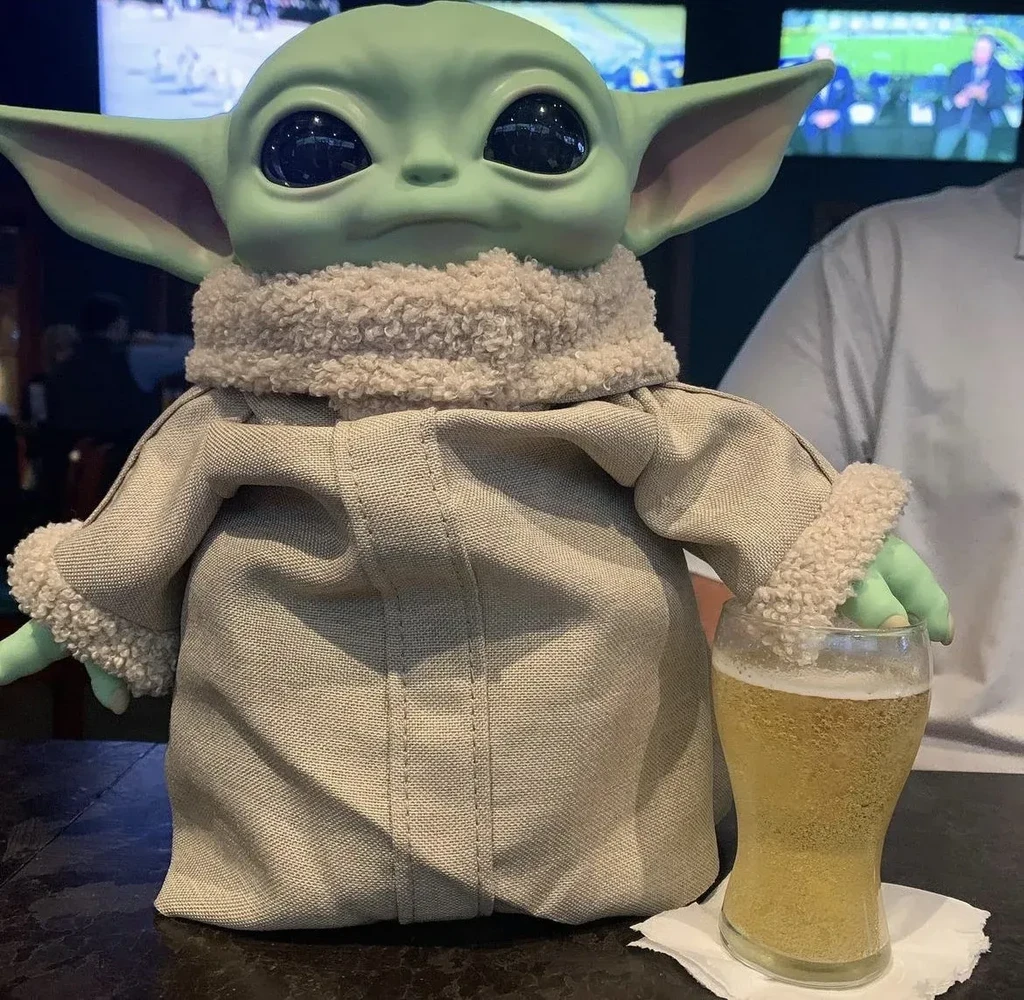 Grogu doll holding a glass of beer on a bar table.