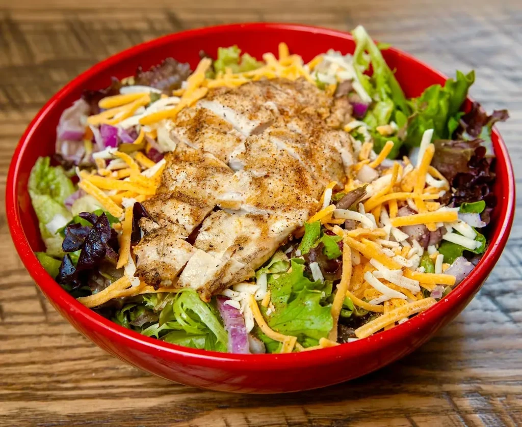 Grilled chicken salad in a red bowl on a wooden table.