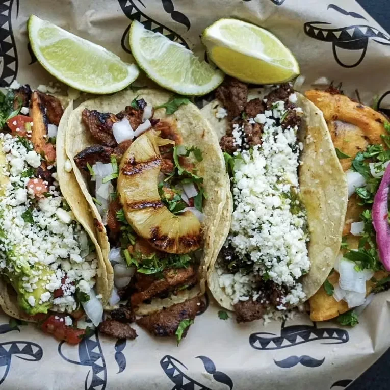Five gourmet tacos with varied fillings, lime, in a themed container.