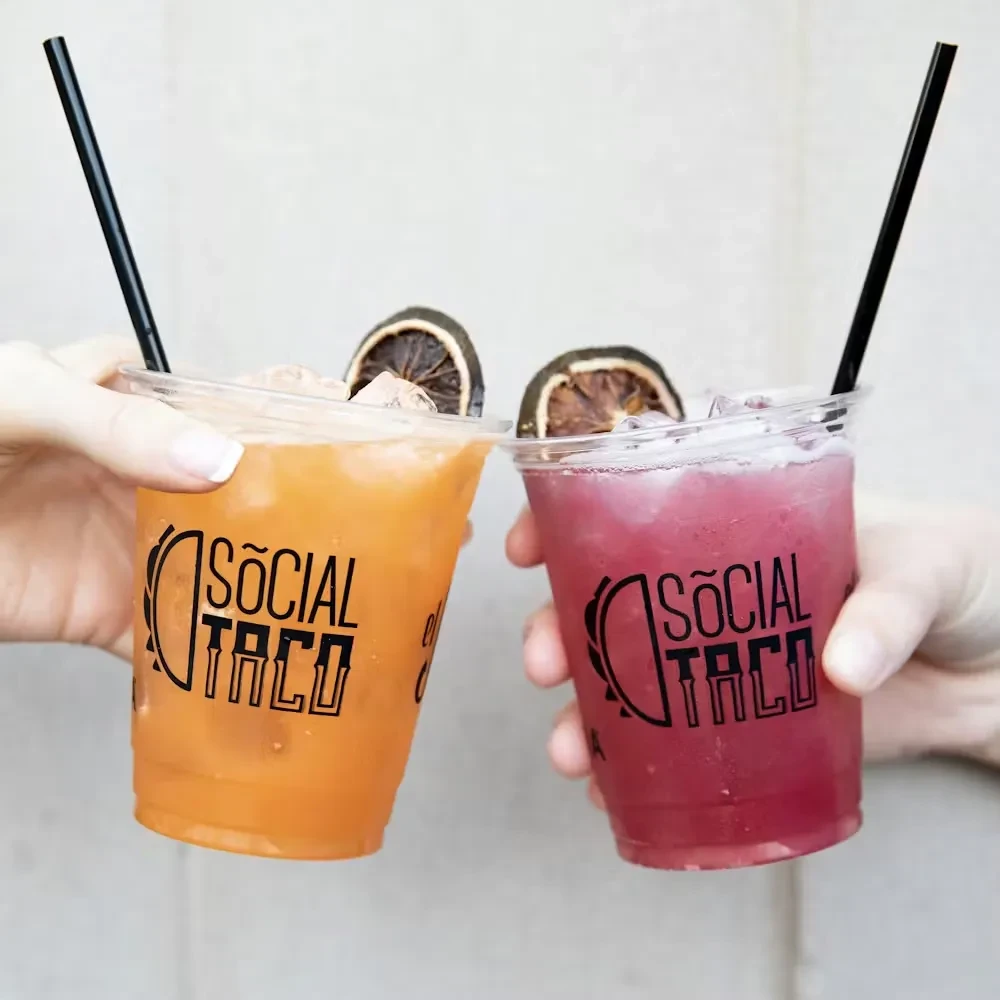 Two hands holding "SOCIAL TACO" cups with orange and pink icy drinks.