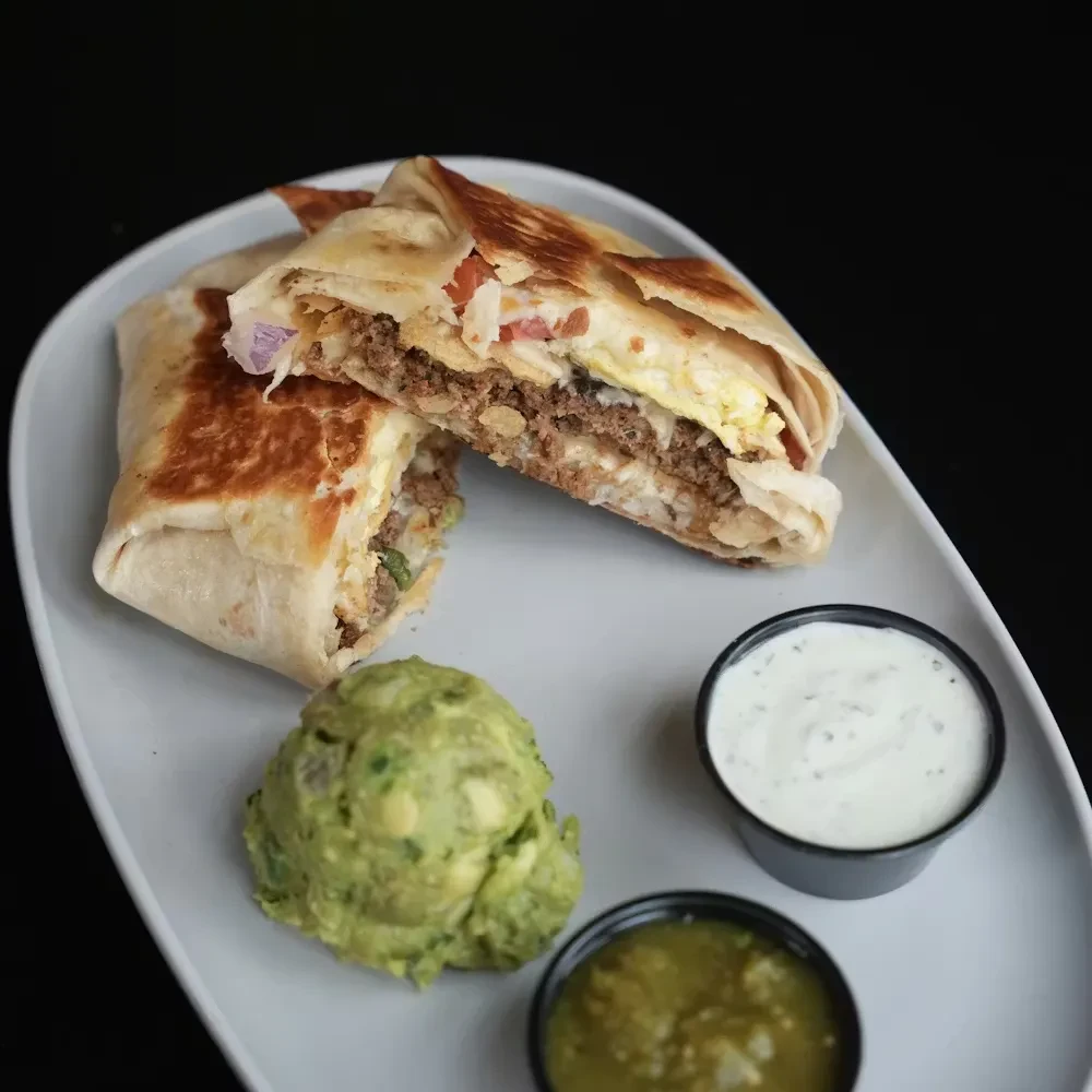 Halved grilled burrito with meat, egg, cheese, guacamole, and two sauces.