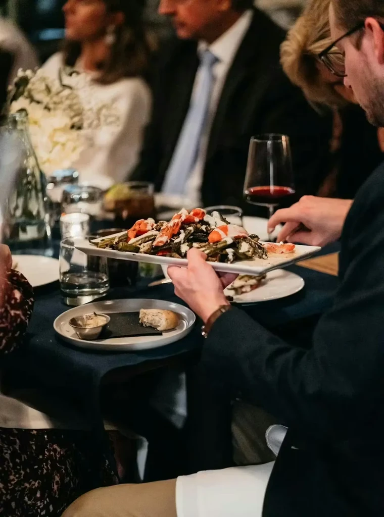 Hands hold platter of roasted vegetables with white sauce at upscale dinner.