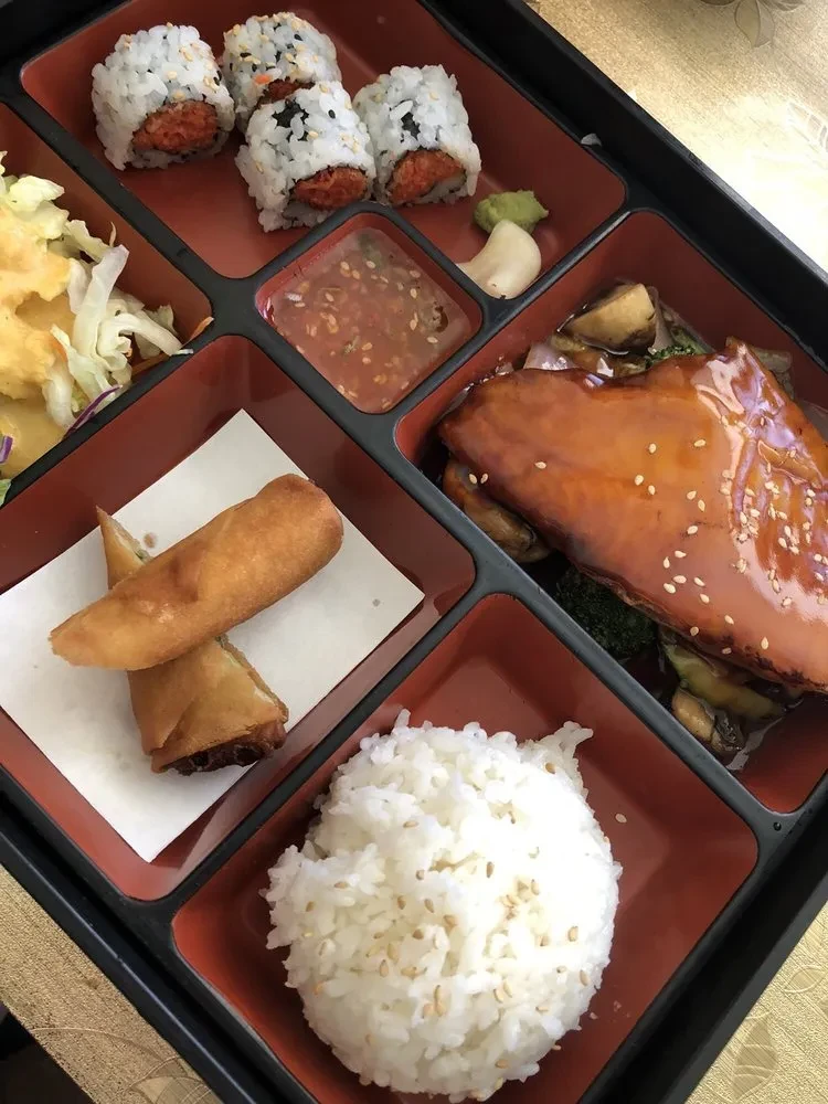 Japanese bento box with sushi, glazed fish, rice, and spring rolls.