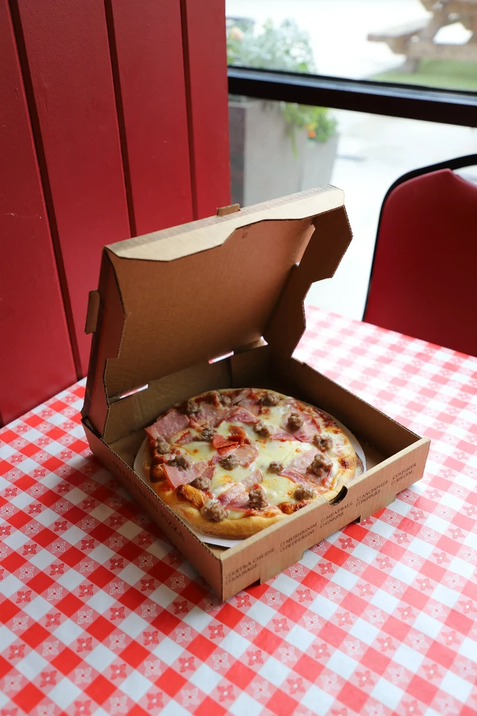 Cooked pizza with meat toppings in box on checkered tablecloth.