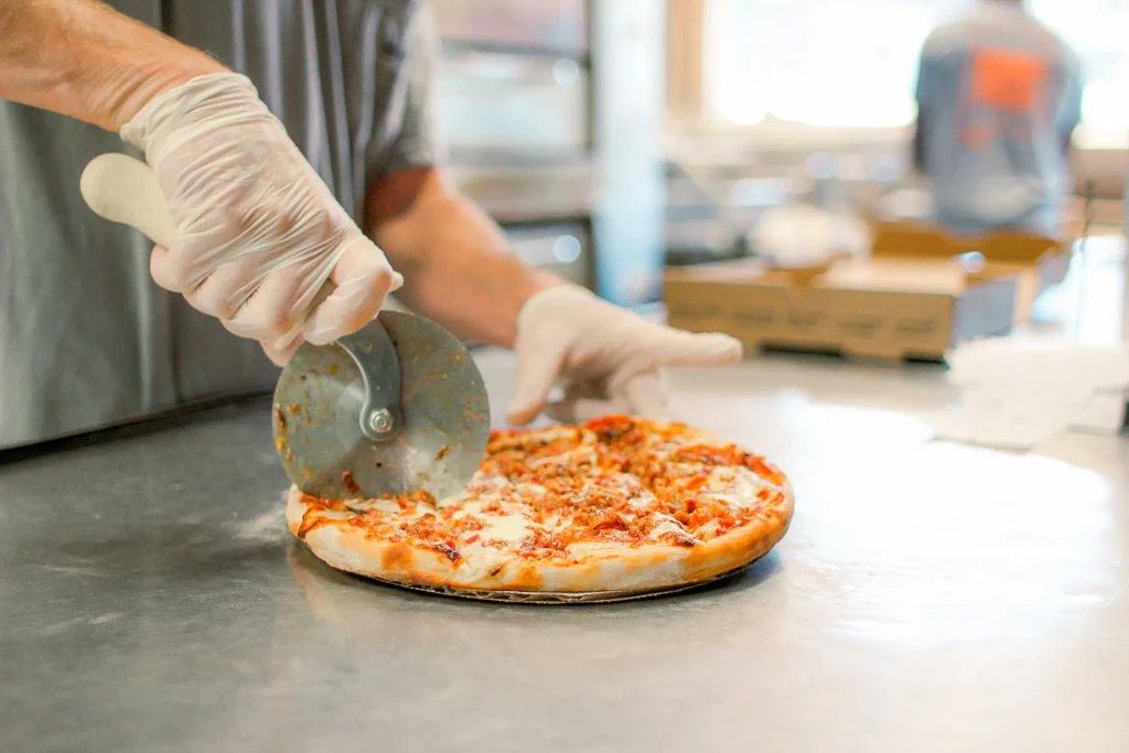Gloved hands cutting a round pizza on a stainless steel counter.