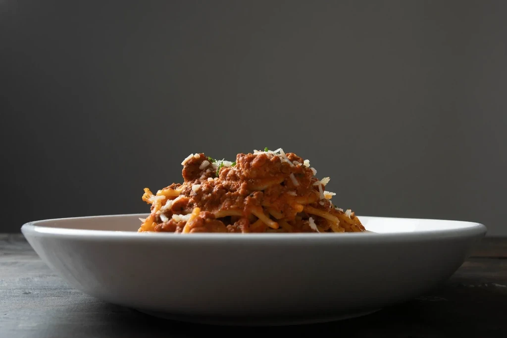 Spaghetti with meat sauce, grated cheese, and herbs in a white bowl.