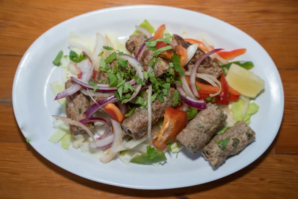 Overhead view of grilled meat kebabs, fresh vegetables, cilantro, and lime.