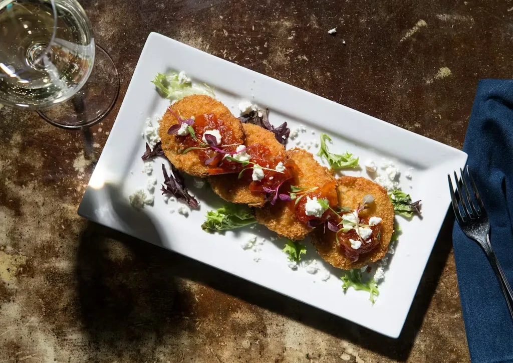 Plated golden-brown fritters with relish, cheese, microgreens, and wine glass.