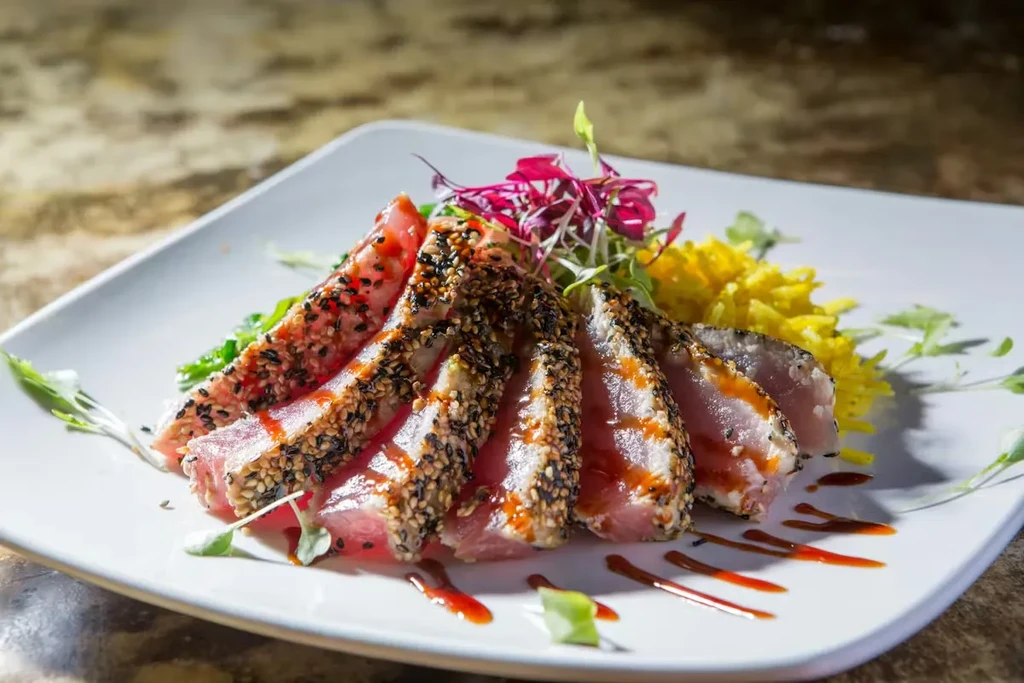 Plated sesame-crusted seared tuna, drizzled sauce, microgreens, yellow side.