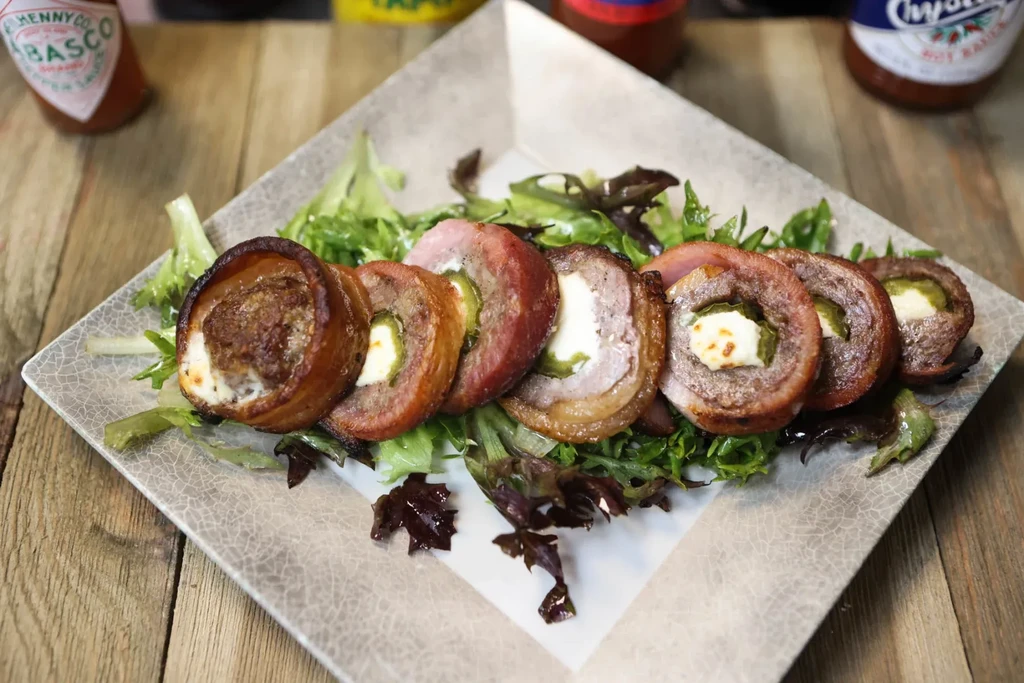 Bacon-wrapped jalapeño poppers with cream cheese on greens, hot sauce nearby.