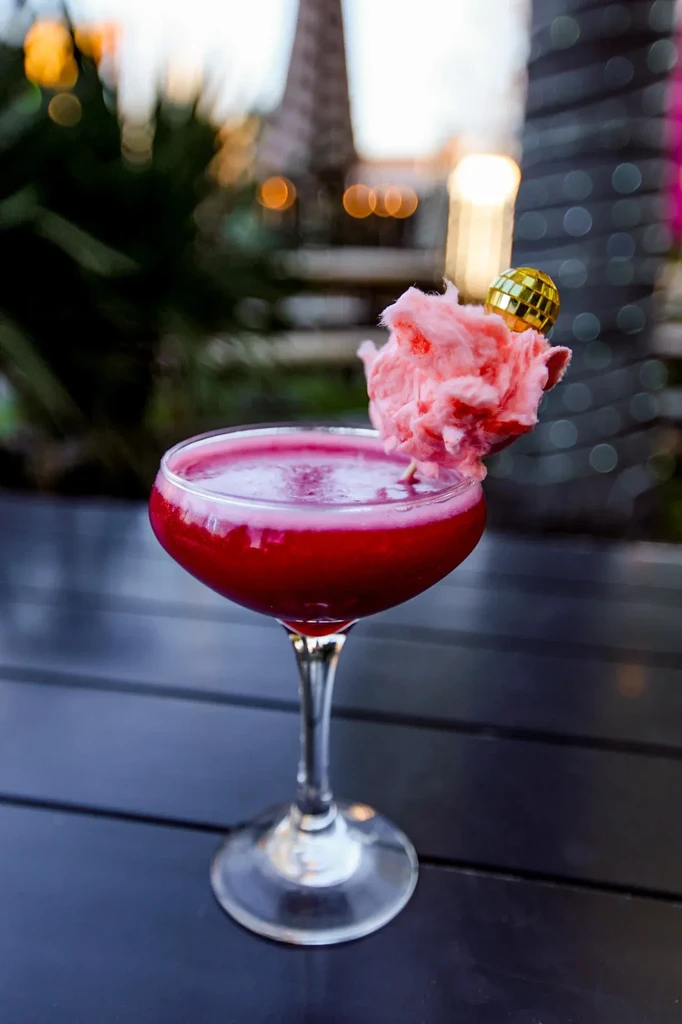 Vibrant red cocktail in coupe glass, garnished with cotton candy and disco ball.
