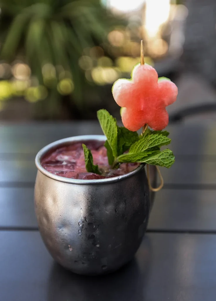 Reddish-purple drink in hammered mug, garnished with mint and watermelon flower.