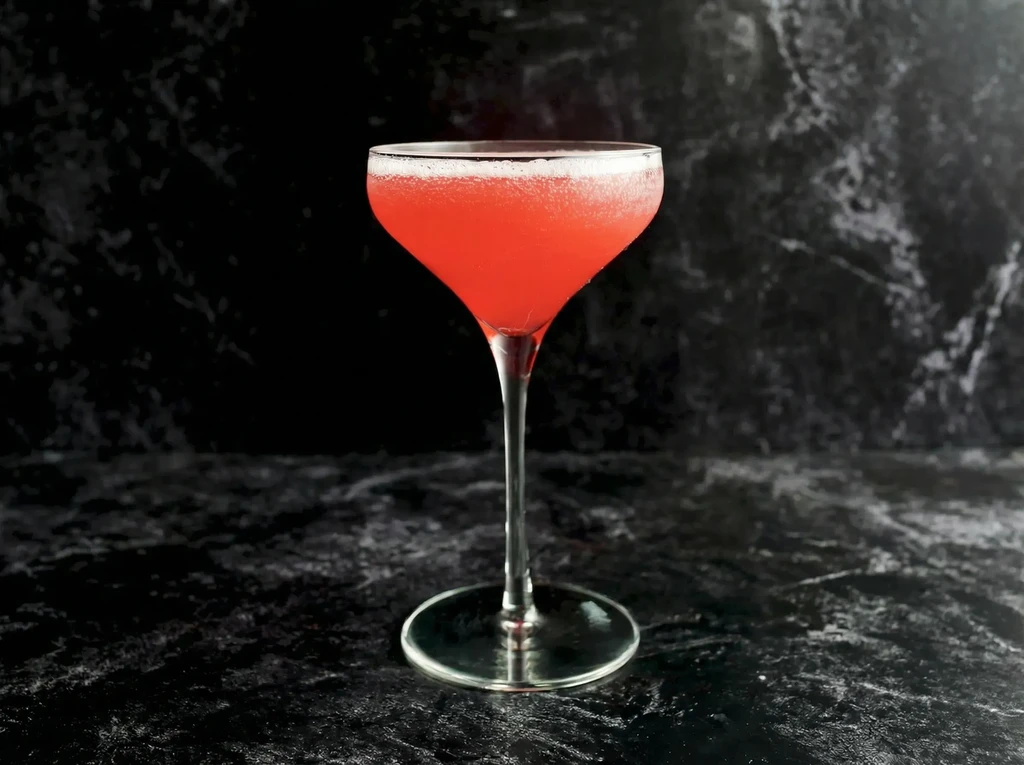 Vibrant red-pink foamy cocktail in a stemmed glass on dark stone.