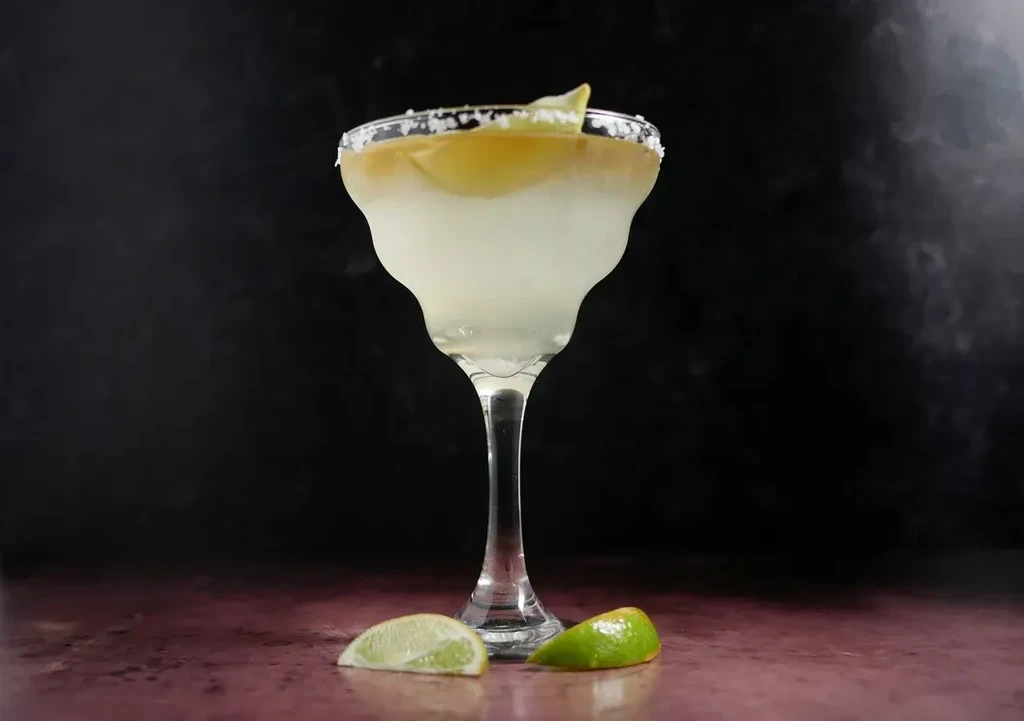 Layered cocktail in margarita glass with salted rim and lime garnishes.