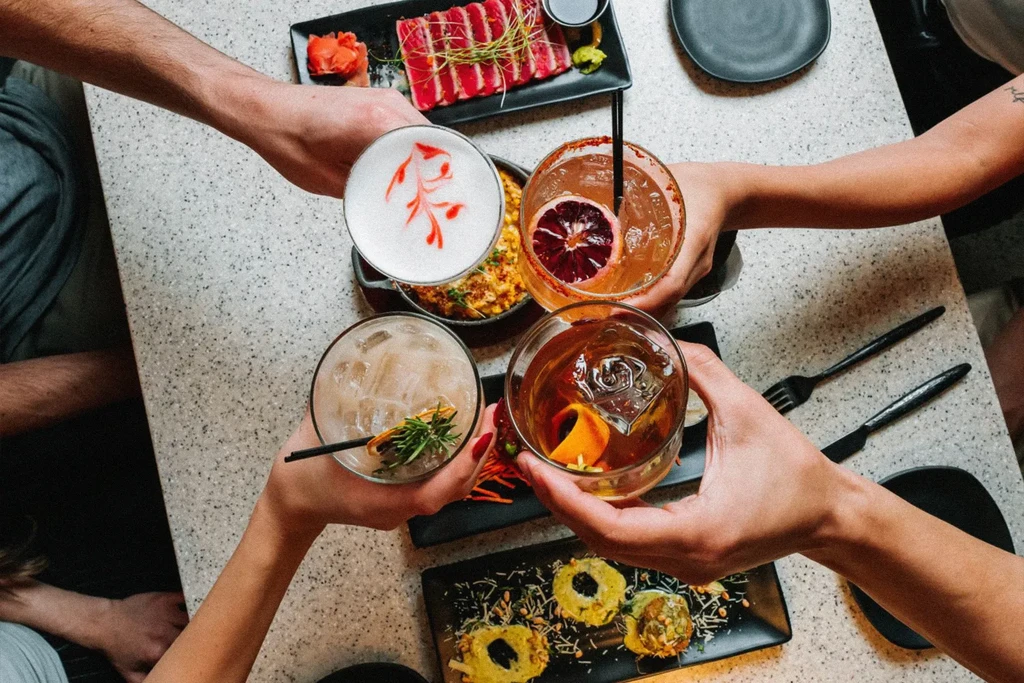 Four hands toast distinct, uniquely garnished cocktails and various artfully presented dishes.