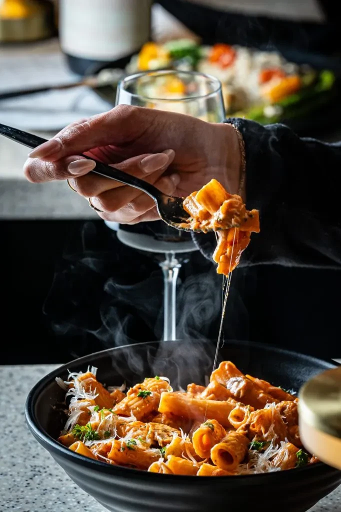 Hand lifts steaming pasta with red sauce, cheese, and herbs.
