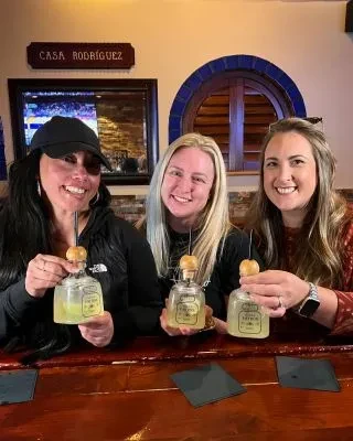Three smiling women at a bar holding unique bottle cocktails.