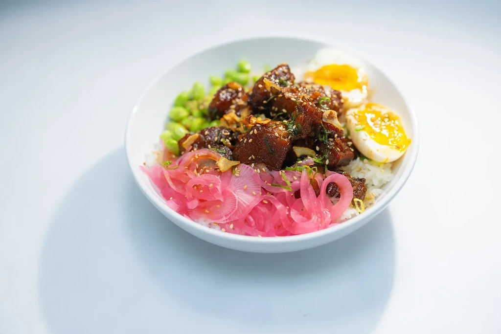 Rice bowl with glazed meat, soft-boiled egg, edamame, and pickled onions.