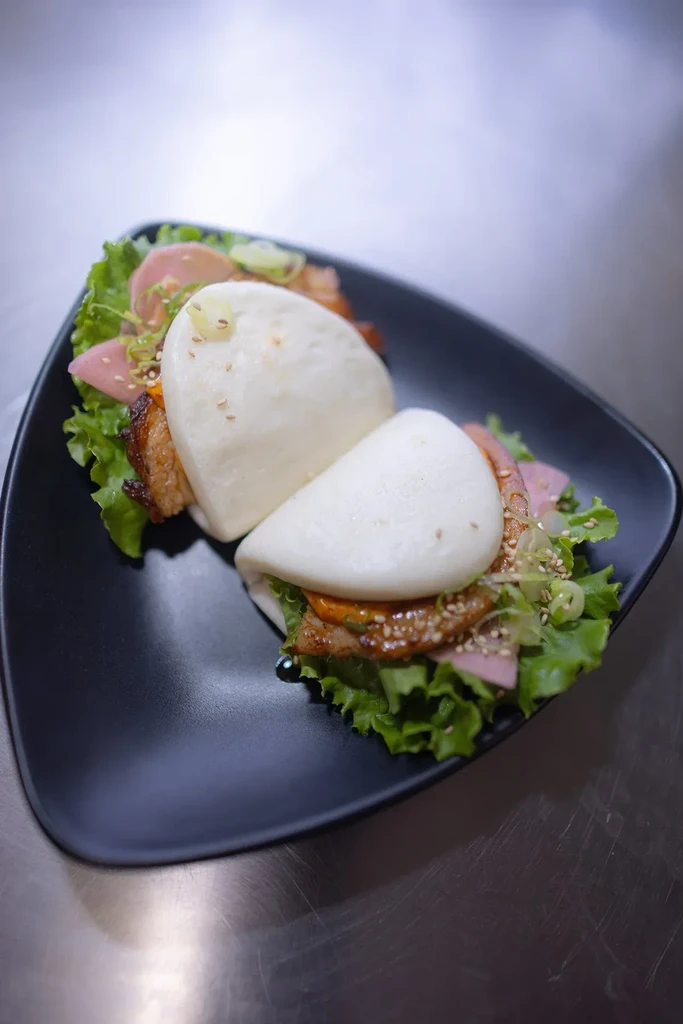 Two steamed bao buns with pork belly, lettuce, radishes on black plate.