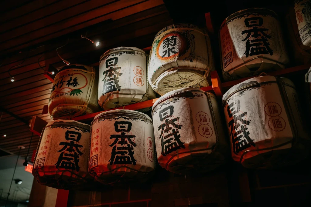 Stacked Japanese sake barrels wrapped in white with calligraphy on red shelf.