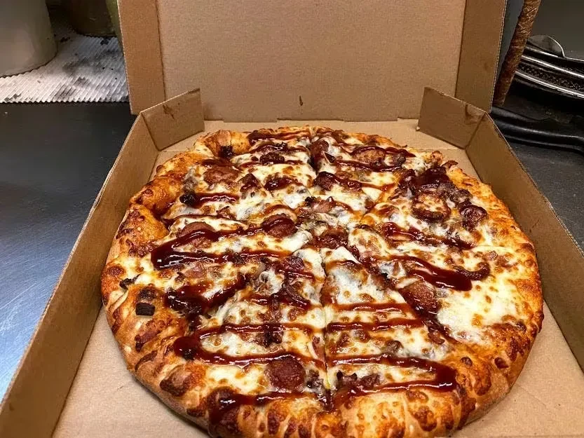 Sliced pizza in open box, with cheese, meats, and BBQ drizzle.