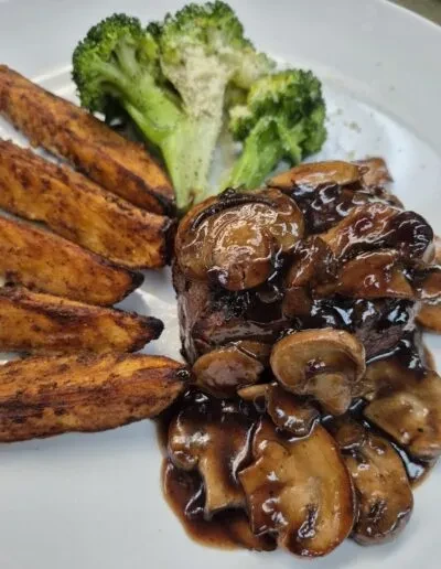 Plated meat with dark mushroom sauce, roasted potatoes, and green broccoli.