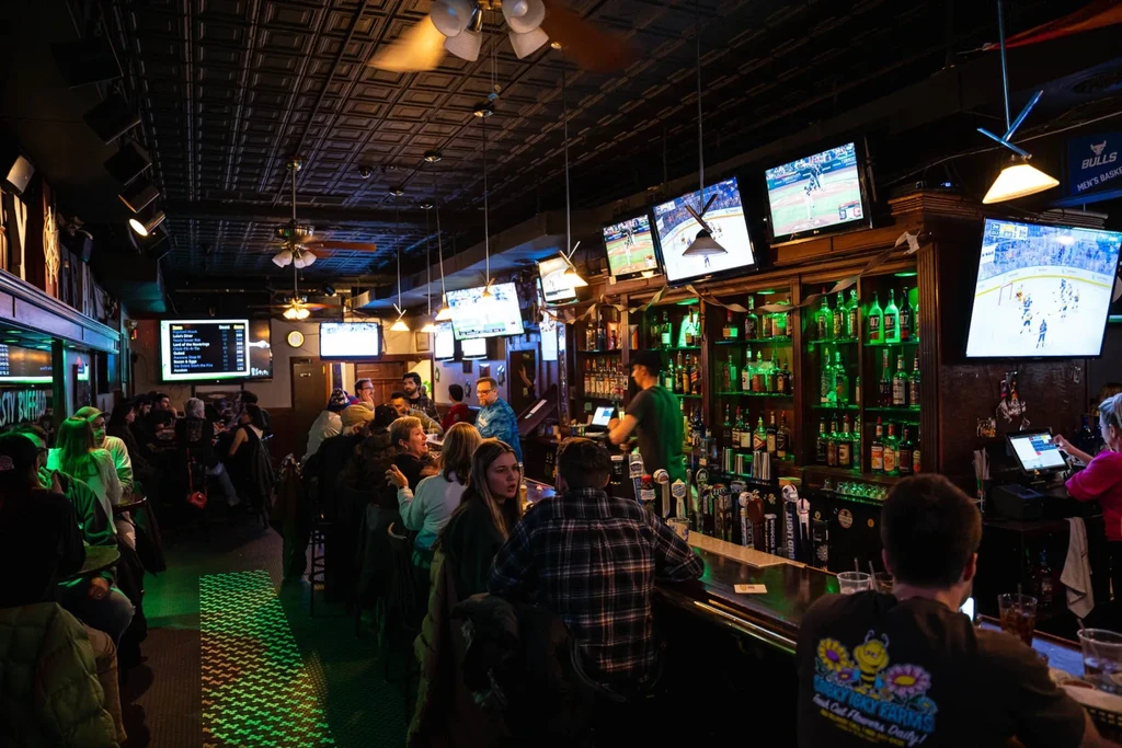 Busy sports bar interior with green-lit bar, sports TVs, and patrons.