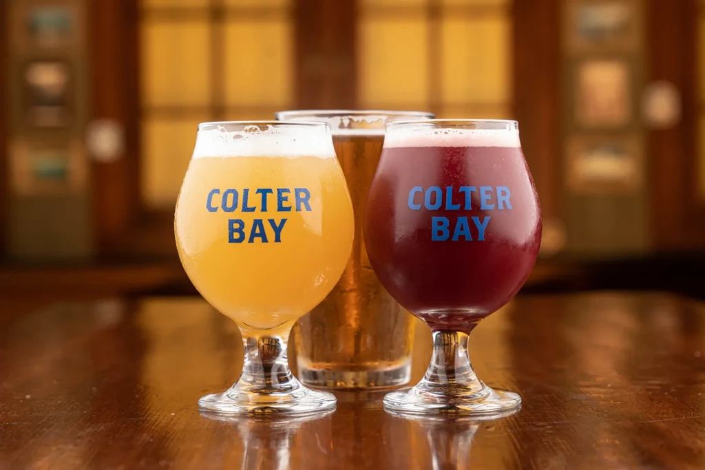 Three glasses of colorful beer, two "COLTER BAY," on a wood surface.