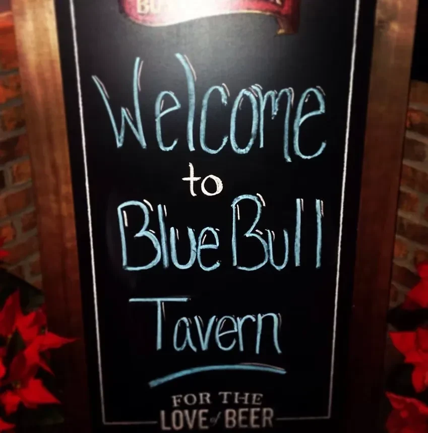 Wooden chalkboard sign reads 'Welcome to Blue Bull Tavern' and beer motto.