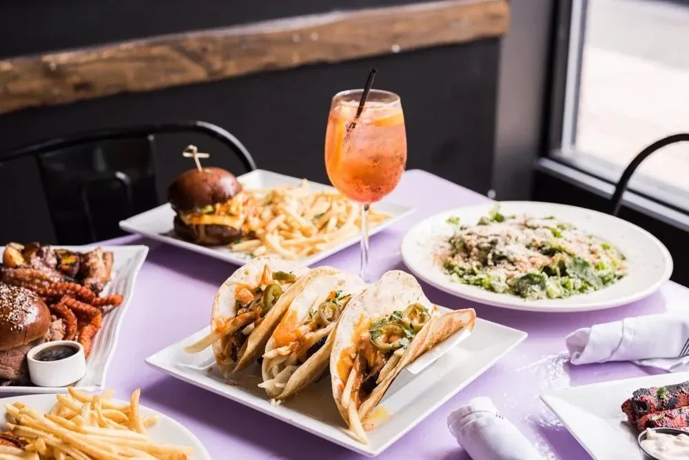 A restaurant table with tacos, burgers, fries, salad, and an orange cocktail.