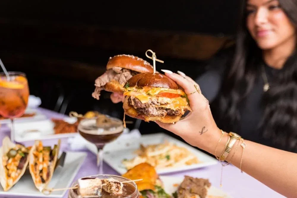Jeweled hand holds large beef burger, other dishes visible.