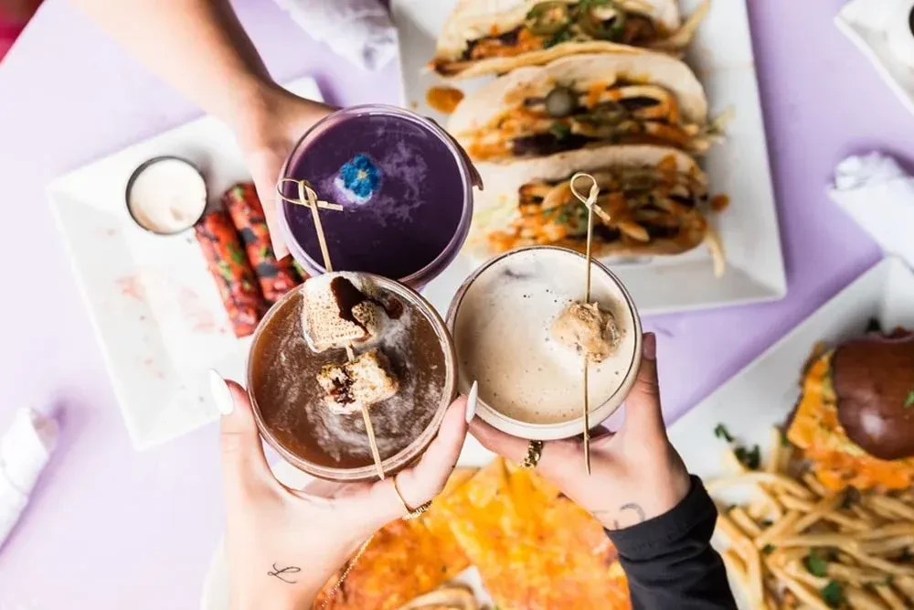 Three hands toast distinct cocktails surrounded by plates of diverse food.