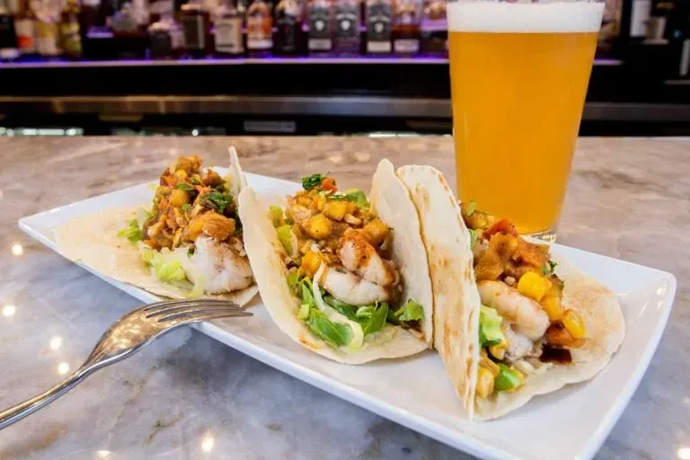 Three shrimp/fish tacos with mango salsa and a beer at a bar.
