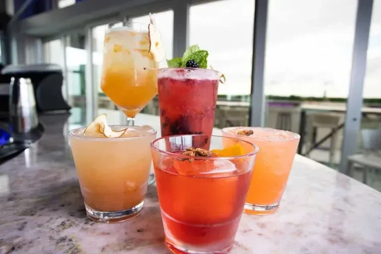 Five colorful, garnished cocktails on a marble counter with an outdoor view.