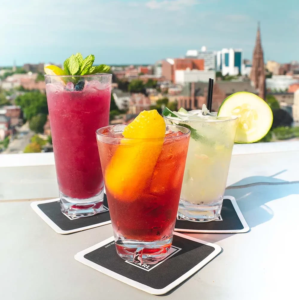 Three colorful, garnished cocktails on an outdoor table with a city skyline.