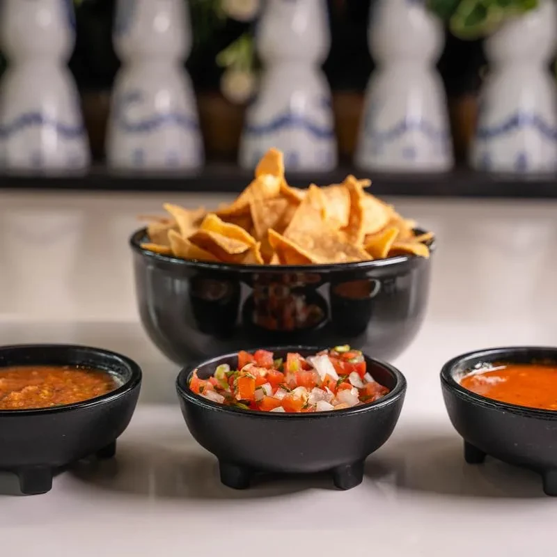 Black bowls with tortilla chips, chunky salsa, pico de gallo, orange sauce.