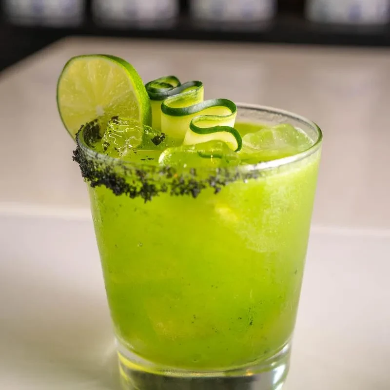 Green cocktail with ice, lime, cucumber, and a dark granular rim.