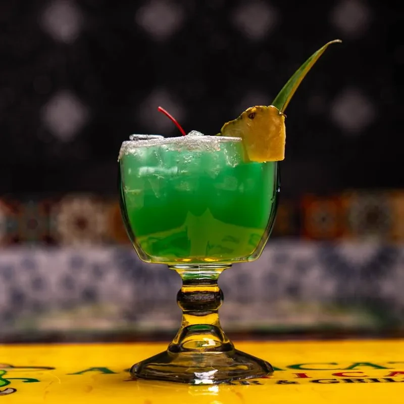 Green cocktail in stemmed glass with sugared rim, cherry, and pineapple.