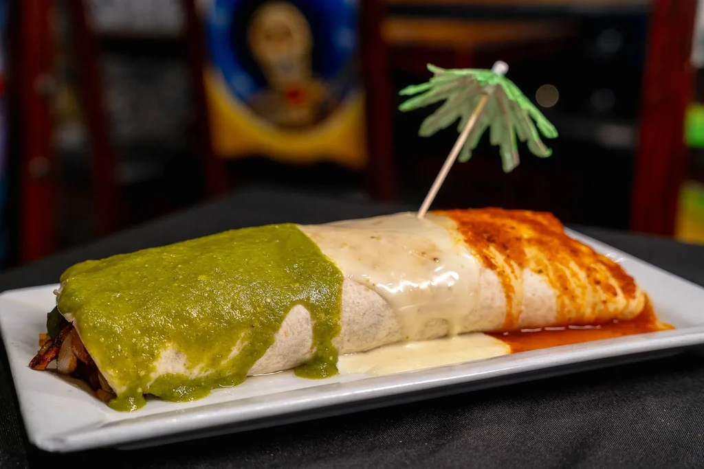 Large burrito with green, white, red sauces and a cocktail umbrella.