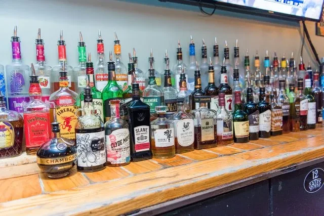 Well-stocked wooden bar counter displays numerous spirit bottles with pour spouts.