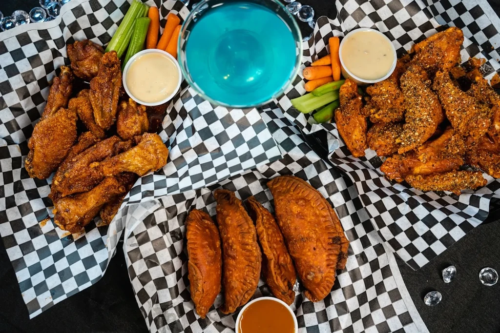 Top-down view of chicken wings, empanadas, vegetables, and a blue drink.