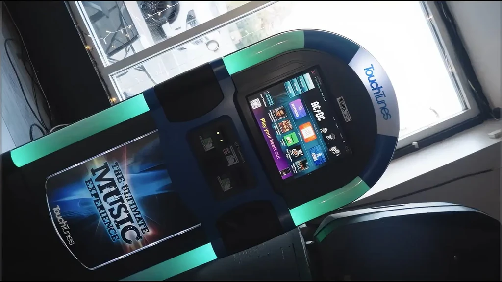 Dark blue Touchtunes jukebox with teal lights, showing album art onscreen.
