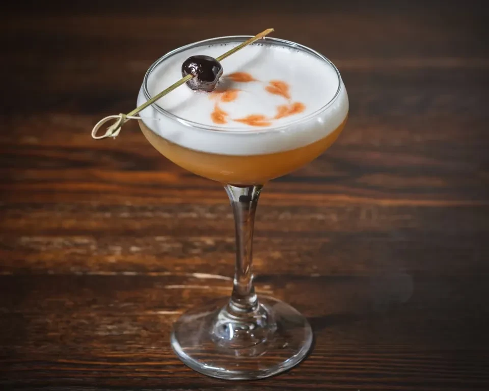 Amber cocktail with patterned foam and cherry on wooden skewer.