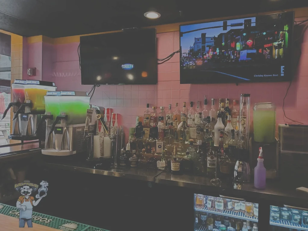 Dimly lit bar with colorful slushie machines, liquor, and pink tiled wall.